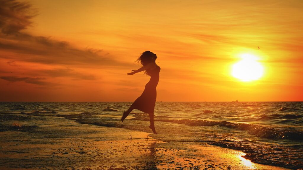 pexels photo 4149949 Side view of anonymous barefoot female jumping with reached arms above sandy shore near wavy ocean under bright cloudy sky at sundown in twilight